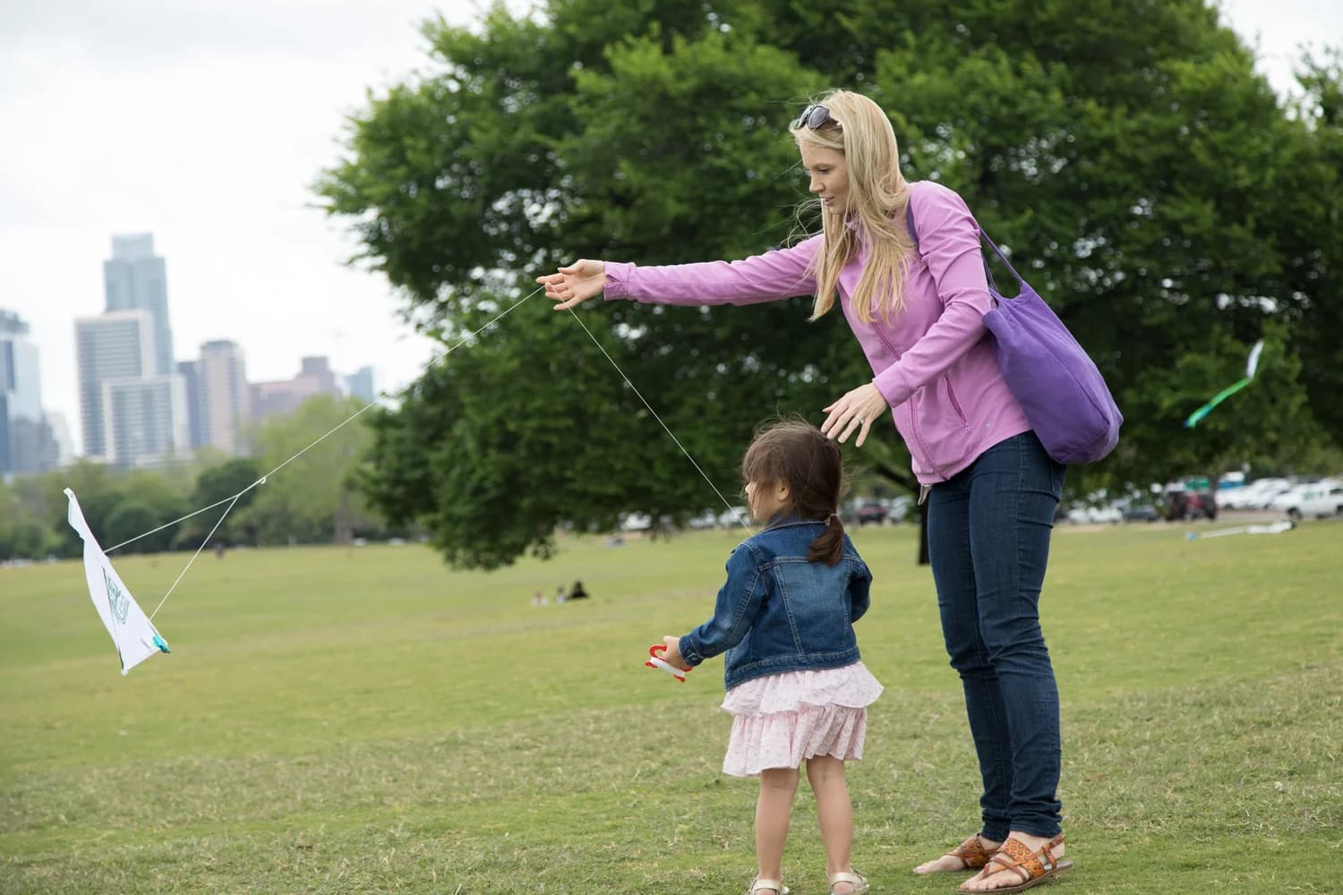 A parent helping a small child fly a kite on the lawn with the skyline behind