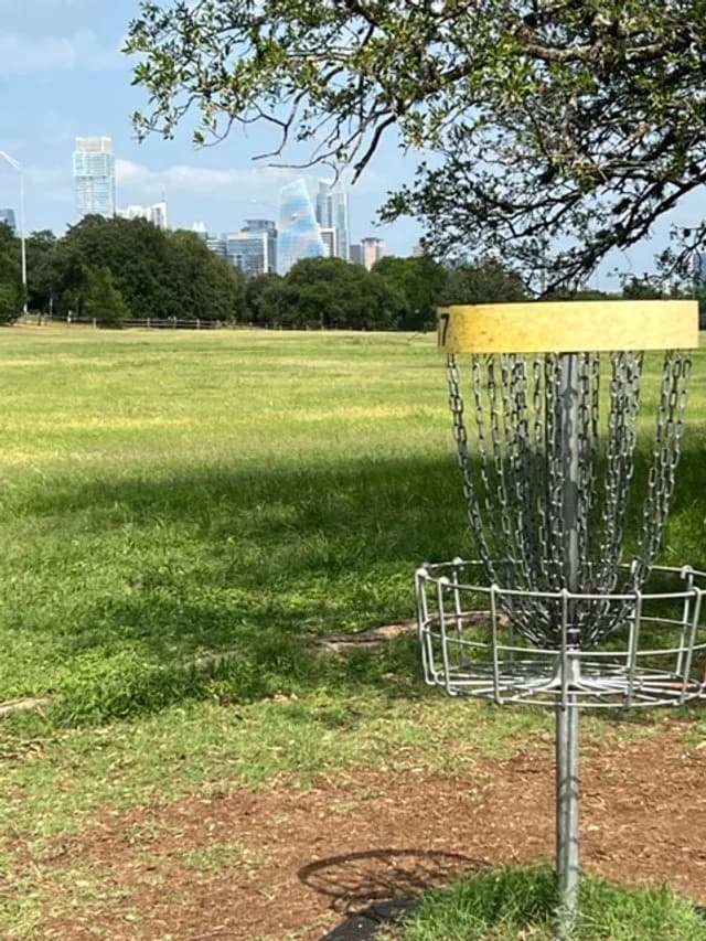 A disc golf basket on the open green of Zilker Park with the Austin skyline in the distance